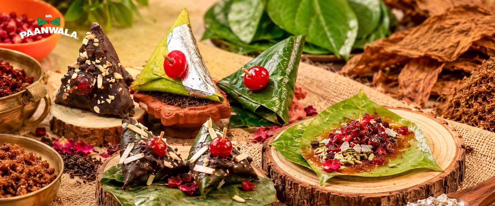 Assorted sweet paan varieties with chocolate, nuts, cherries, and colourful fillings arranged on wooden plates with paan ingredients.